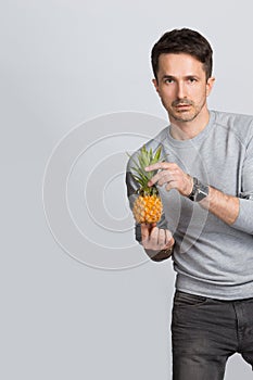 Handsome young man holding a small yellow pineapple in his hands and looking at the camera on a gray background.