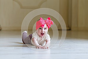 Cute happy 6 months baby girl with bright bow crawling indoor. Pretty smiling baby girl with open mounth. Light