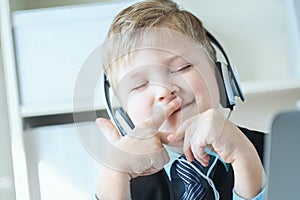 Cute happy 6 year old boy in suit listening to music or audio tutorial on headphones at the office background.