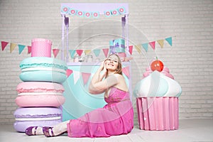 Cute happy adult girl posing in fashionable dress among huge fabulous sweets and cakes