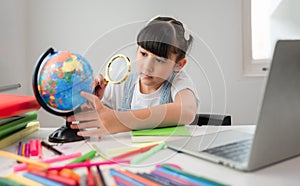 Cute happy Asian child girl student sitting at a desk with a laptop and using a magnifying glass to look and search the map and