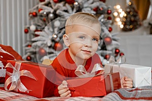 Cute happy boy in red sweater lying on bed and dreaming about christmas present box on holiday morning in beautiful room interior