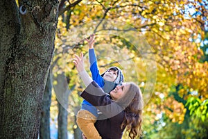 Cute, happy, boy smiling and hugging with his mom among yellow leaves. Little child having fun with mother in autumn park. Concept