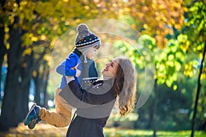 Cute, happy, boy smiling and hugging with his mom among yellow leaves. Little child having fun with mother in autumn park. Concept