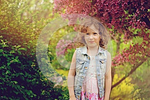 Cute happy child girl in jeans vest enjoying spring near blooming crab apple tree in country garden