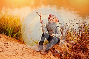 Cute happy child girl playing with stick on autumn river side