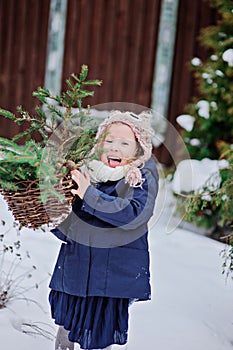 Cute happy child girl playing in winter snowy garden with basket of fir branches