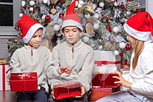 Cute happy excited children, boy and girl in santa hats unwrap christmas present box in beautiful room.