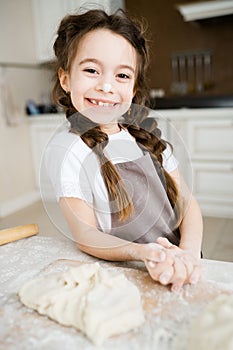 A cute happy girl with pigtails and an apron leans on the table with flour and dough and looks at the camera smiling