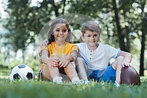 cute happy kids with soccer and rugby balls sitting on grass and smiling