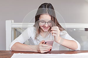 Cute happy laughing tween girl in glasses looking at smartphone in her hands talking on video call with smile.