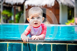 A cute and happy little baby holds to the side of the tropical pool and looks to the camera with interest. Infant girl swim at