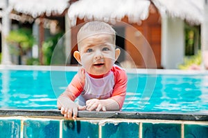 A cute and happy little baby holds to the side of the tropical pool and looks to the camera and laught. Infant girl in swim suit