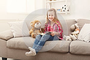 Happy little female child hugging her teddy bear and reading book on sofa at home