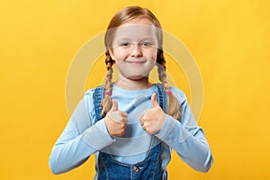 Cute happy little girl shows thumb up with two hands. A child with pigtails on a yellow background