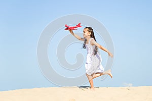 Cute happy little girl wearing a white dress running on the sandy beach by the sea and playing with the red t