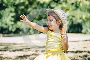 Cute happy little girl in yellow dress and hat eating fruit ice cream. Summer fun child playing in park. Summertime vibrant color