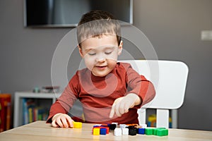 A cute happy little toddler boy of two years old sits at a children\'s table and plays with multi-colored blocks