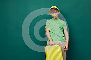 a cute, happy man stands on a green background, dressed in a yellow panama hat, holding a bright travel suitcase in his