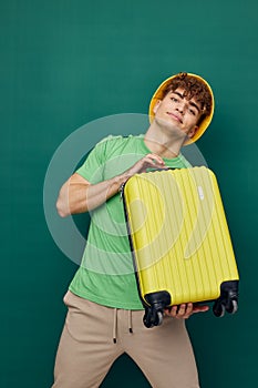 a cute, happy man stands on a green background, dressed in a yellow panama hat, holding a bright travel suitcase in his