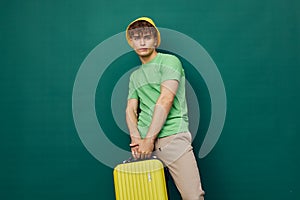 a cute, happy man stands on a green background, dressed in a yellow panama hat, holding a bright travel suitcase in his