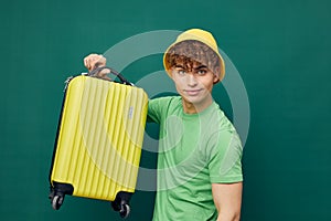 a cute, happy man stands on a green background, dressed in a yellow panama hat, holding a bright travel suitcase in his