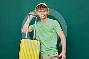 a cute, happy man stands on a green background, dressed in a yellow panama hat, holding a bright travel suitcase in his