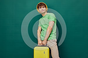 a cute, happy man stands on a green background, dressed in a yellow panama hat, holding a bright travel suitcase in his