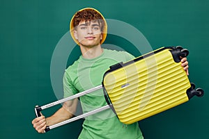 a cute, happy man stands on a green background, dressed in a yellow panama hat, holding a bright travel suitcase in his