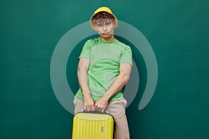 a cute, happy man stands on a green background, dressed in a yellow panama hat, holding a bright travel suitcase in his