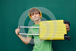 a cute, happy man stands on a green background, dressed in a yellow panama hat, holding a bright travel suitcase in his