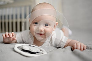 Cute happy newborn baby lying on bed on his stomach next to rattle. Portrait of an infant in a bodysuit, resting at home