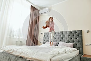 A cute happy small girl is having fun jumping on a big bed with her teddy bear
