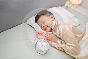 Cute happy smiling little Asian boy kid in pajama taking a nap, sleeping in bed with alarm clock