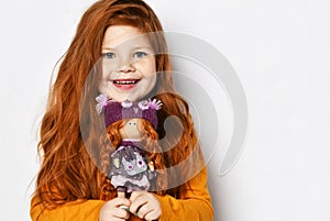 Cute happy smiling red-haired kid girl in orange sweatshirt holds small redhair doll her portrait in hands at face