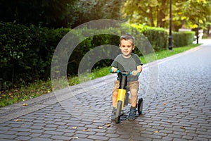 A cute happy toddler boy of two or three years old rides a bicycle or balance bike in a city park on a sunny summer day.