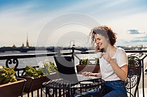 Cute happy young girl working on computer and smiling. Beautiful woman with laptop on a balcony with a landscape on the river Neva