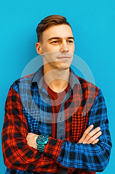 Cute happy young man with a stylish hairstyle in a stylish T-shirt in a fashionable checkered colorful shirt posing near the blue