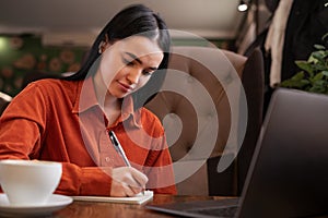 Cute hardworking girl making notes, copying online information , using laptop while working or studying, sitting in cafe