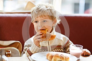 Cute healthy kid boy eating croissant and drinking strawberry milkshake in cafe. Happy child having breakfast with