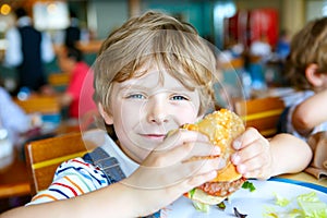 Cute healthy preschool boy eats hamburger sitting in cafe outdoors