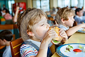 Cute healthy preschool boy eats hamburger sitting in school canteen