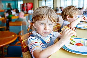 Cute healthy preschool boy eats hamburger sitting in school canteen