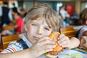 Cute healthy preschool boy eats hamburger sitting in school canteen