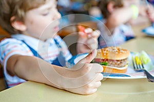 Cute healthy preschool boy eats hamburger sitting in school canteen