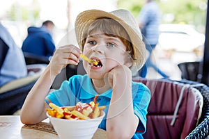 Cute healthy preschool kid boy eats french fries potatoes with ketchup sitting in cafe outdoors. Happy child eating