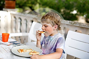 Cute healthy preschool kid boy eats fresh pizza sitting on terrace in summer, outdoors