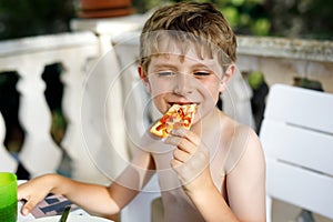 Cute healthy preschool kid boy eats fresh pizza sitting on terrace in summer, outdoors