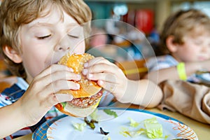 Cute healthy preschool kid boy eats hamburger sitting in school or nursery cafe. Happy child eating healthy organic and