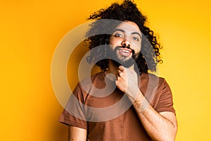 Cute hindu man with afro hairstyle touches his beard with his hand and looks at the camera smiling
